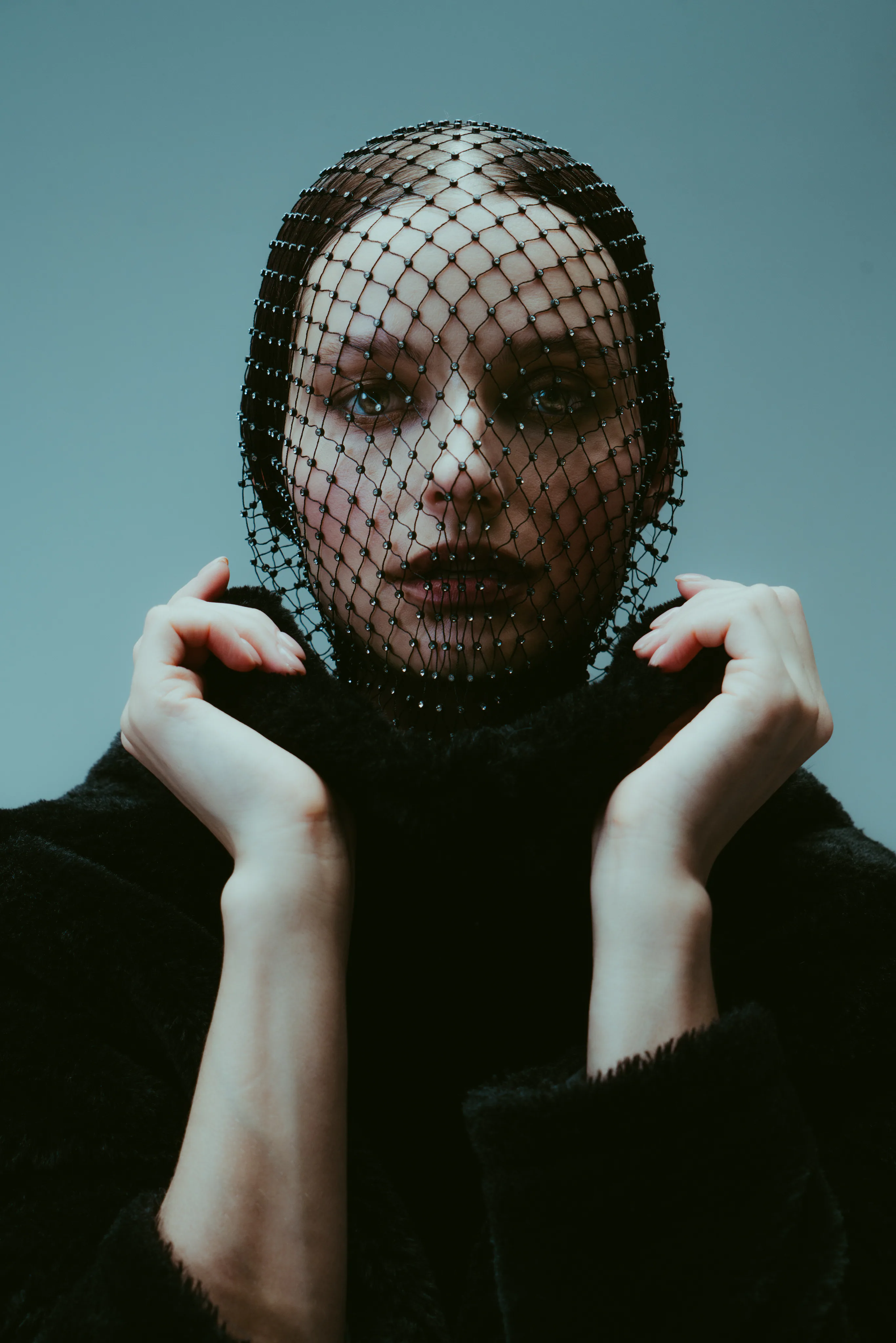 A woman in a black fishnet hood and black coat holding up her collar against a blue-grey backdrop